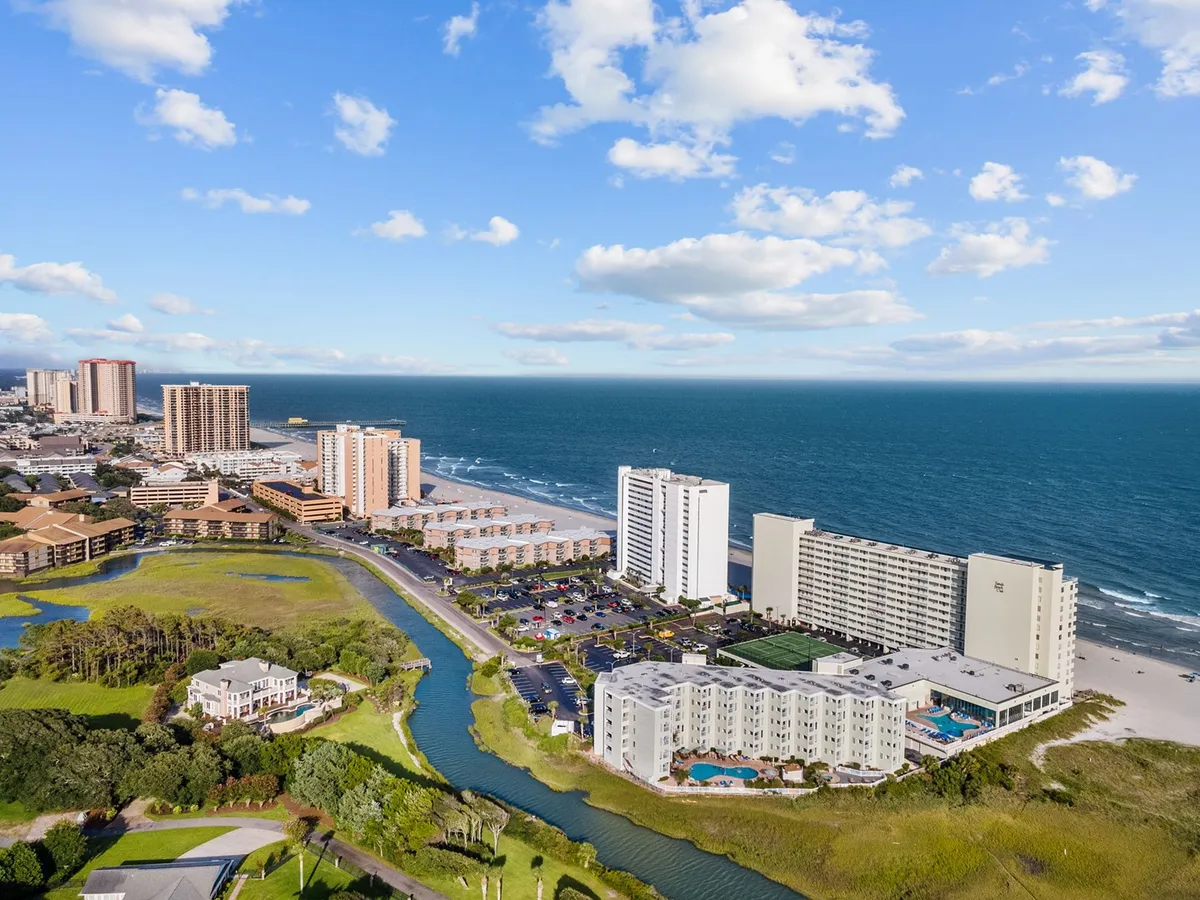 This image offers a sweeping view of the ocean, beachfront buildings, lush green landscape, and a nearby canal, all set under a vibrant sky. The scene highlights the lively beachfront location and conveys the sense of easy access to beach activities and nearby attractions—key themes in the section.