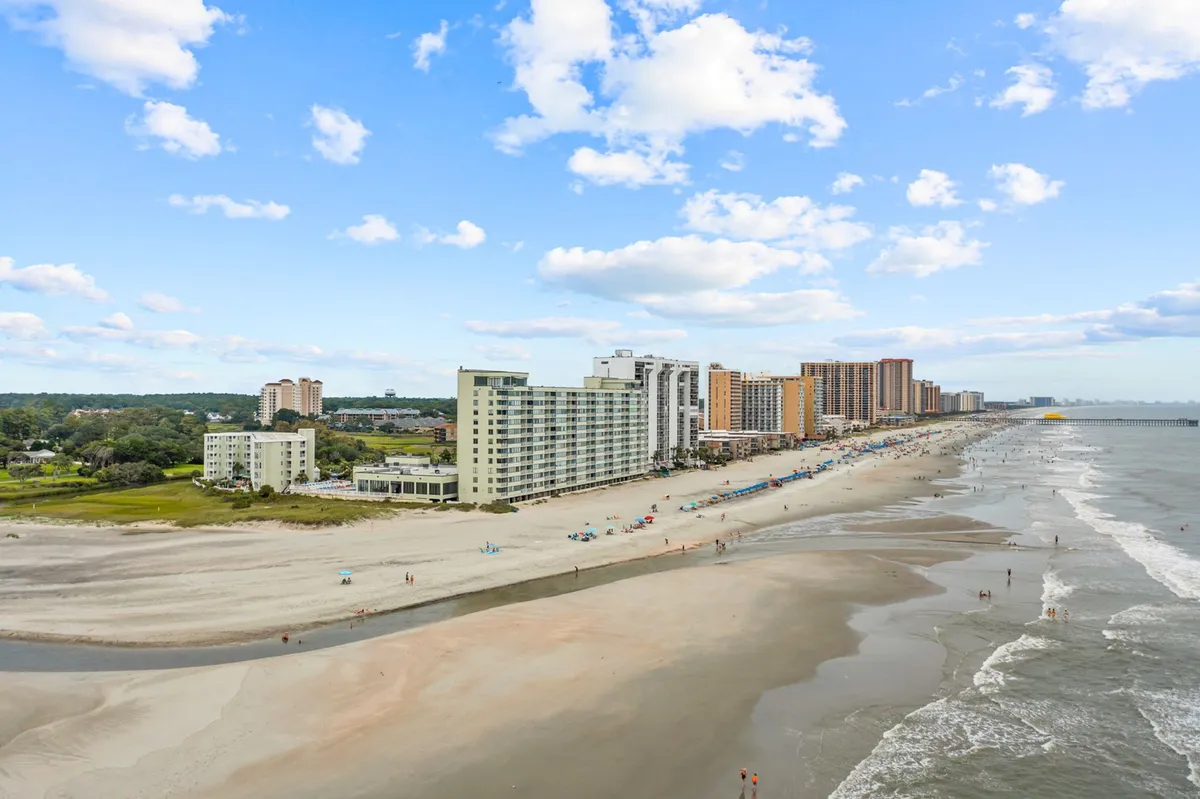 This exterior image captures the beach, ocean, adjacent buildings, and a dramatic sky, with people visible sunbathing, swimming, and playing on the sand. The atmosphere is relaxed and carefree, encapsulating the fun, social aspect of a Myrtle Beach group getaway.