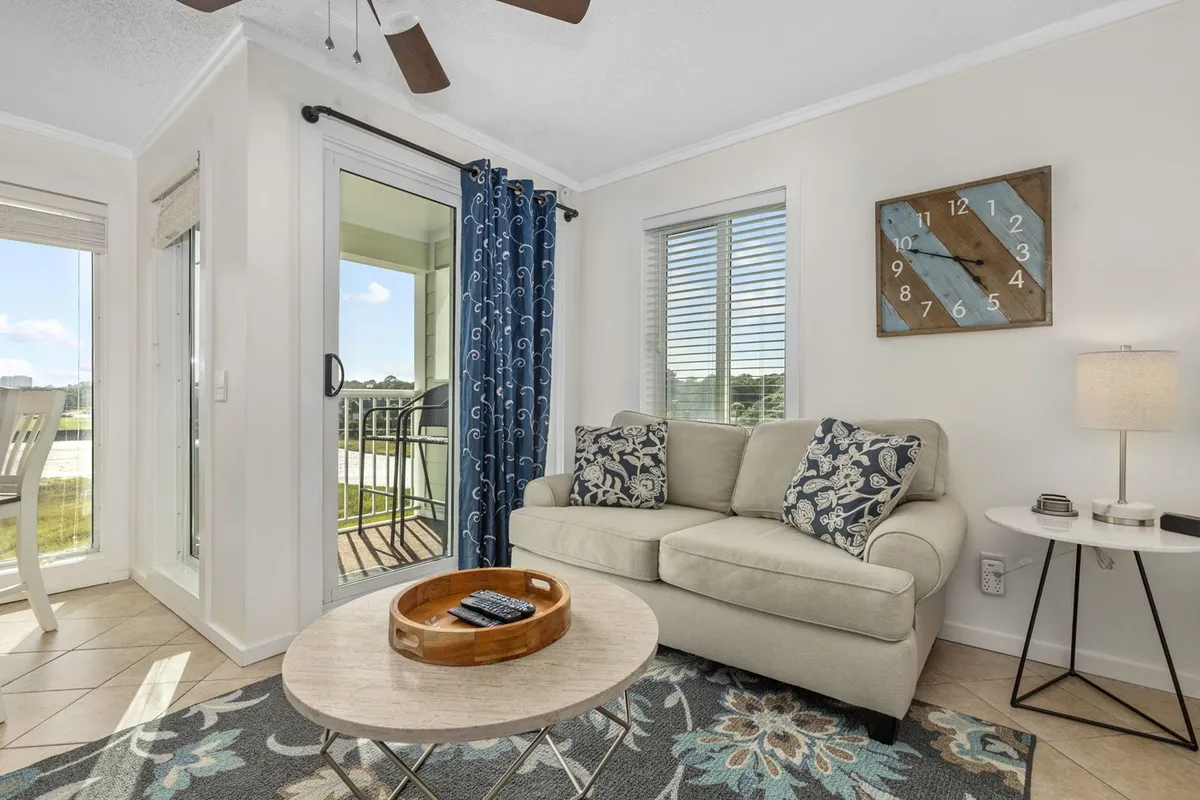 A plush sofa and coffee table face a door opening directly onto the balcony, with curtains framing the view and a decorative clock adding a homey touch. The space invites relaxation, reading, and taking in the ocean atmosphere.