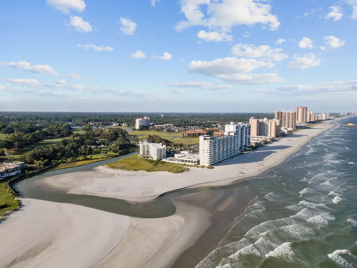 This image captures the beach shoreline with soft ocean waves, high-rise buildings in the background, and lush greenery, evoking the peaceful, coveted section of Shore Drive mentioned in the section. The relaxed beach scene aligns with the promise of tranquil escapes and relaxing strolls.
