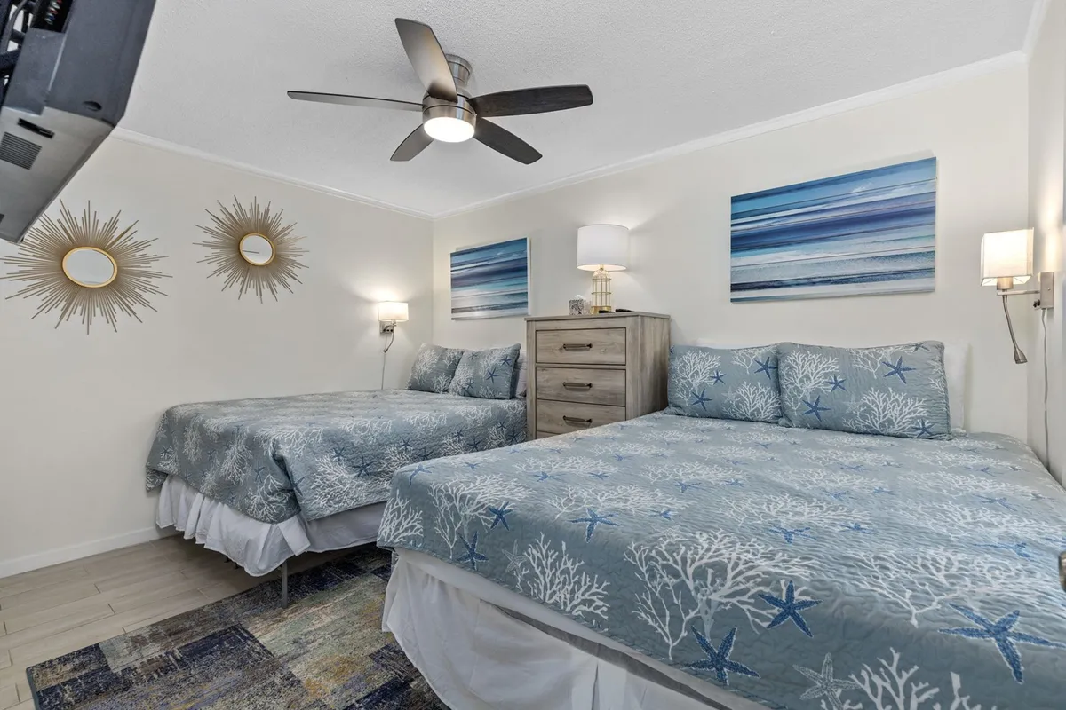 The photo features a serene bedroom with two beds adorned in coral-patterned bedding, a wooden dresser, wall art of ocean waves, and decorative mirrors with a sunburst design. The room feels bright, airy, and designed for comfort, perfect for families or groups.