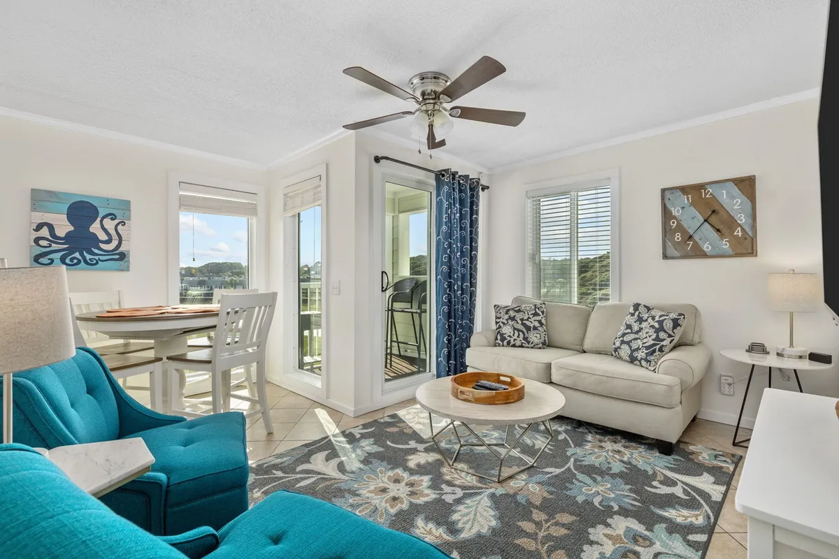 The image shows a sunlit, inviting living area with vibrant turquoise chairs, a stylish coffee table adorned with decorative accents, and tasteful artwork on the wall, creating a cozy yet modern atmosphere perfect for relaxing or socializing.