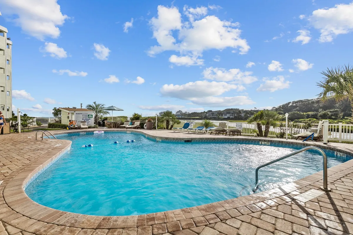 An inviting outdoor pool surrounded by lounge chairs and lush greenery, with the Sands Beach Club building visible in the background. The scene exudes a cheerful, social atmosphere perfect for relaxation or making summer memories.