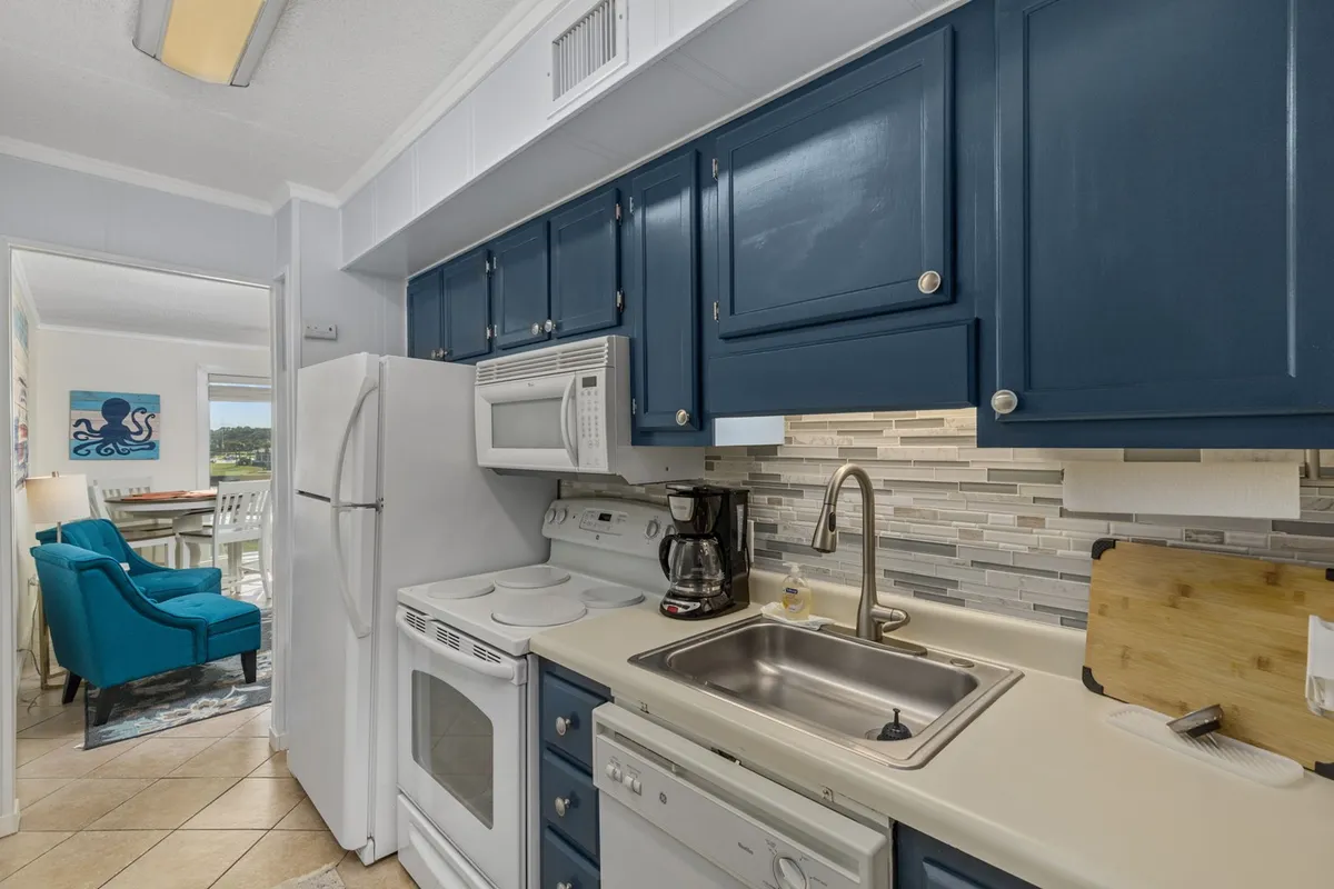 This image shows a lively kitchen scene with sleek blue cabinetry, stainless steel appliances including a full-size fridge and stove, and a modern backsplash. The space is lit with cheerful natural light, emphasizing its open and inviting atmosphere.
