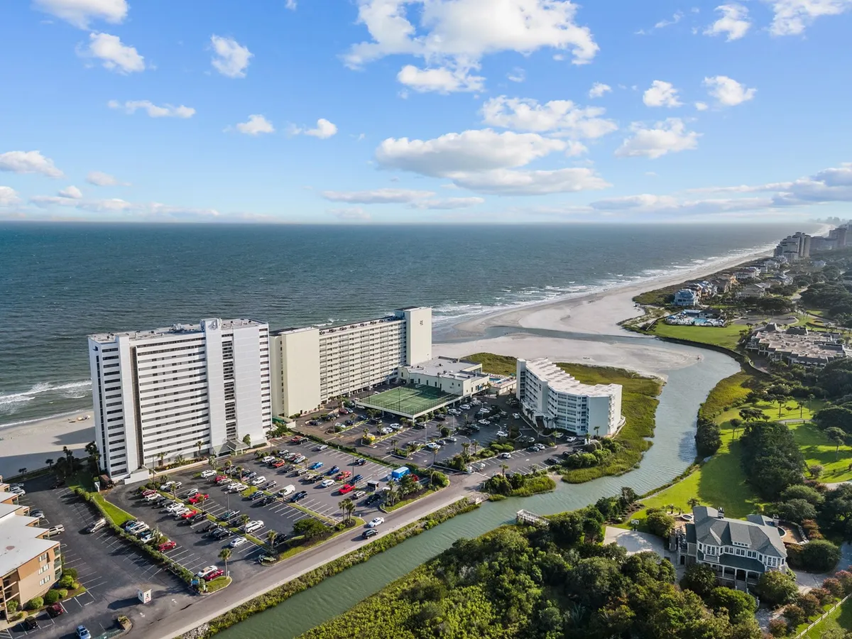 A panoramic view of the ocean and beach is framed by beachfront buildings and a parking area. The setting is lively, inviting visitors to participate in beach activities, water sports, and explore nearby attractions.