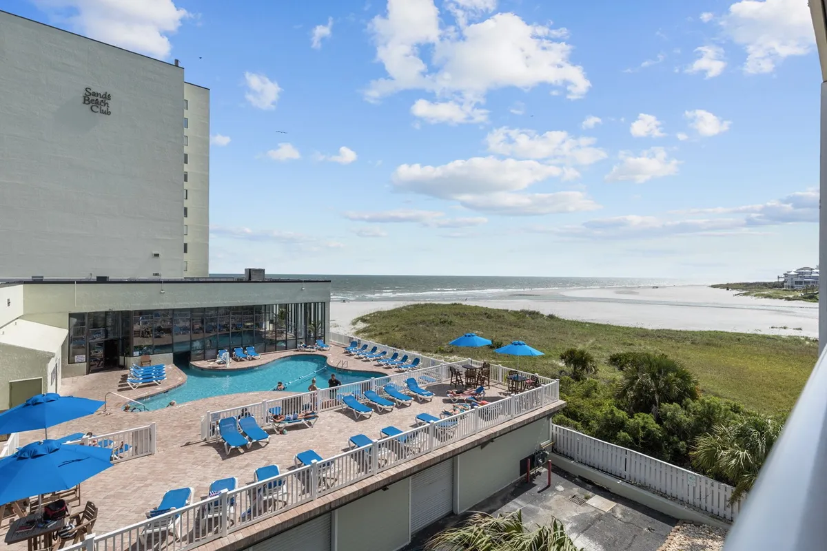 A sparkling swimming pool overlooking the sandy beach and ocean, dotted with lounge chairs and umbrellas, creating a classic beach resort vibe.