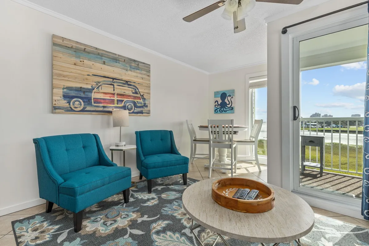 A welcoming open-plan living space featuring teal armchairs, a wooden coffee table, a dining table set for meals, and a large sliding glass door that floods the area with sunlight and provides seamless access to an ocean-facing balcony.