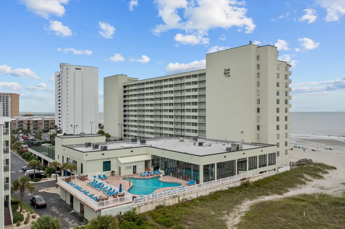 The image highlights a stunning pool area with clear blue water, the adjacent sandy beach, and a welcoming hotel building. The sky is bright, and the overall mood suggests relaxation and vacation enjoyment, with easy access to both swimming and the ocean.