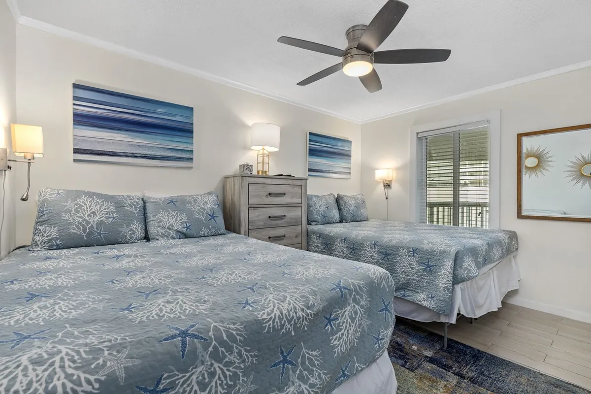 This image showcases two beds with blue coral-patterned bedding against a wall with abstract ocean-themed artwork. A modern dresser and a window letting in light create a cozy, inviting, and restful setting.